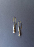 The Elodie Pearl Earrings by LLUME Jewelry feature gold threaders with dangling rows of white pearls, elegantly displayed on a dark grey background—perfect bridal earrings for wedding jewelry.