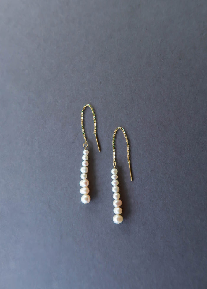 The Elodie Pearl Earrings by LLUME Jewelry feature gold threaders with dangling rows of white pearls, elegantly displayed on a dark grey background—perfect bridal earrings for wedding jewelry.