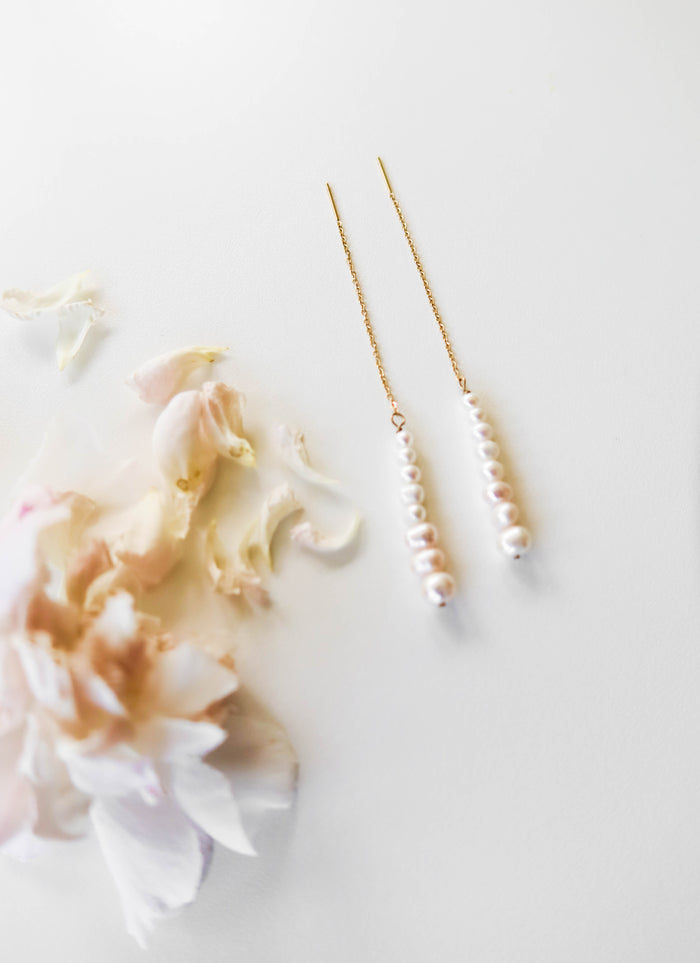 The Elodie Pearl Earrings by LLUME Jewelry feature a line of small white pearls on delicate threaders, elegantly displayed on a white surface beside pale pink flower petals—perfect as wedding jewelry.