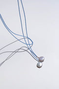 Two Esme Lariat Necklaces by LLUME Jewelry, each featuring a delicate blue cord and a single round white pearl pendant, rest on a white surface with soft shadows—an elegant choice for timeless style lovers.