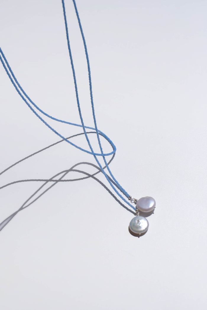 Two Esme Lariat Necklaces by LLUME Jewelry, each featuring a delicate blue cord and a single round white pearl pendant, rest on a white surface with soft shadows—an elegant choice for timeless style lovers.