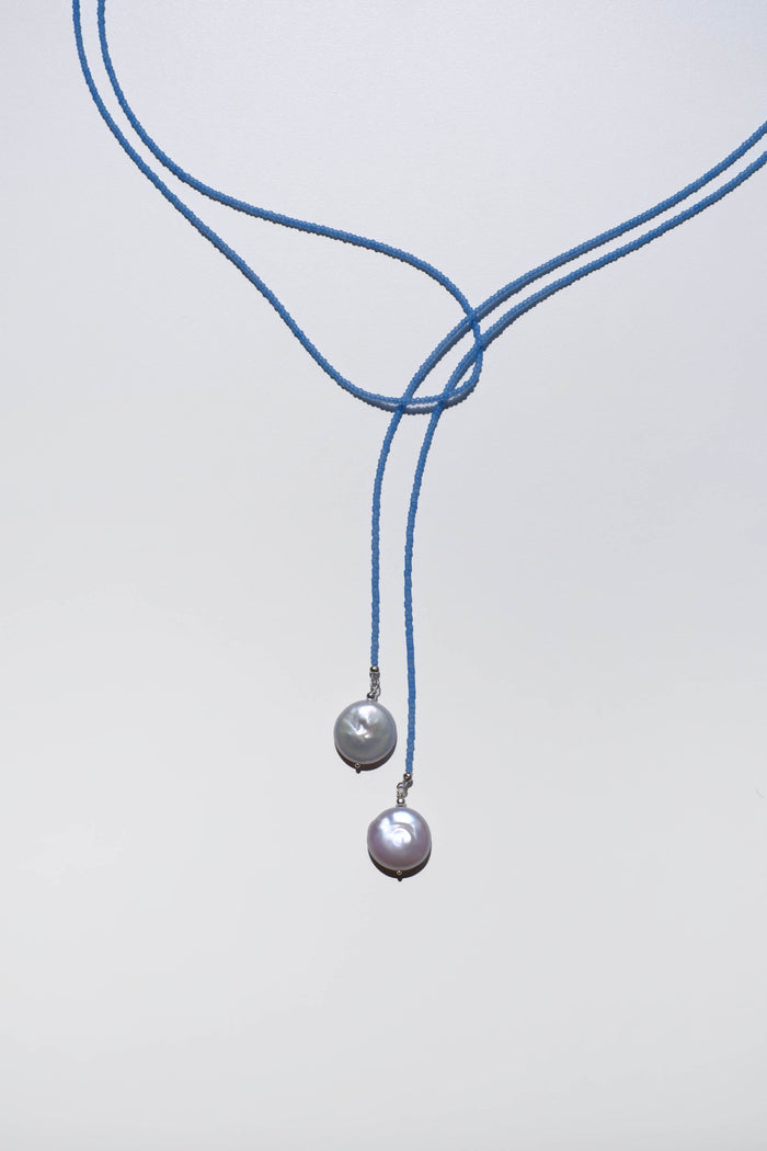 The Esme Lariat Necklace by LLUME Jewelry features a blue cord with two baroque pearls hanging at varying lengths, offering an elegant accent against a plain light background.