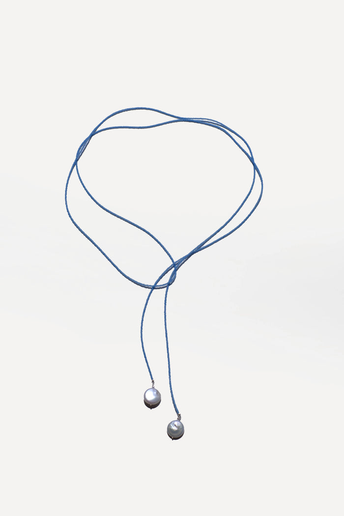 The Esme Lariat Necklace by LLUME Jewelry features a blue cord with silver bead accents, styled in a heart-like shape on a white background—a timeless accessory to elevate any look.