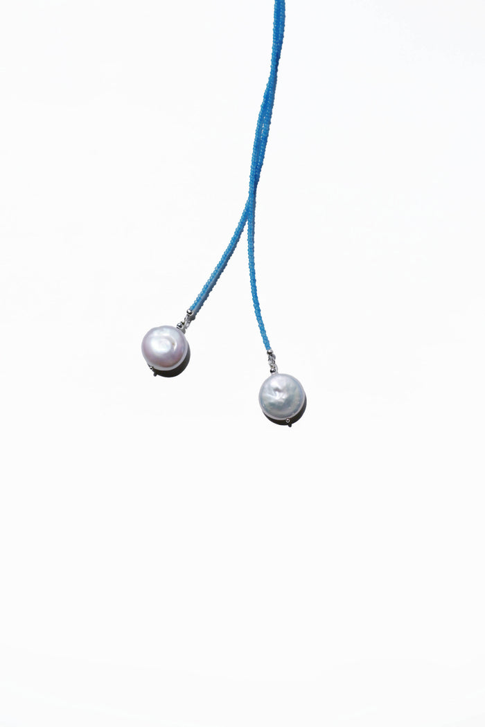 Two blue strings, each tipped with a round white pearl, hang against a plain white background. This showcases the Esme Lariat Necklace from LLUME Jewelry—a timeless, minimalistic piece of elegance.