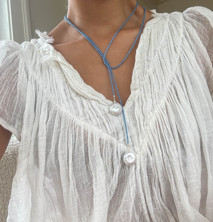 A person wearing a sheer, white, crinkled v-neck blouse styled with the Esme Lariat Necklace by LLUME Jewelry—an elegant blue beaded necklace featuring two large white pearls at varying lengths.