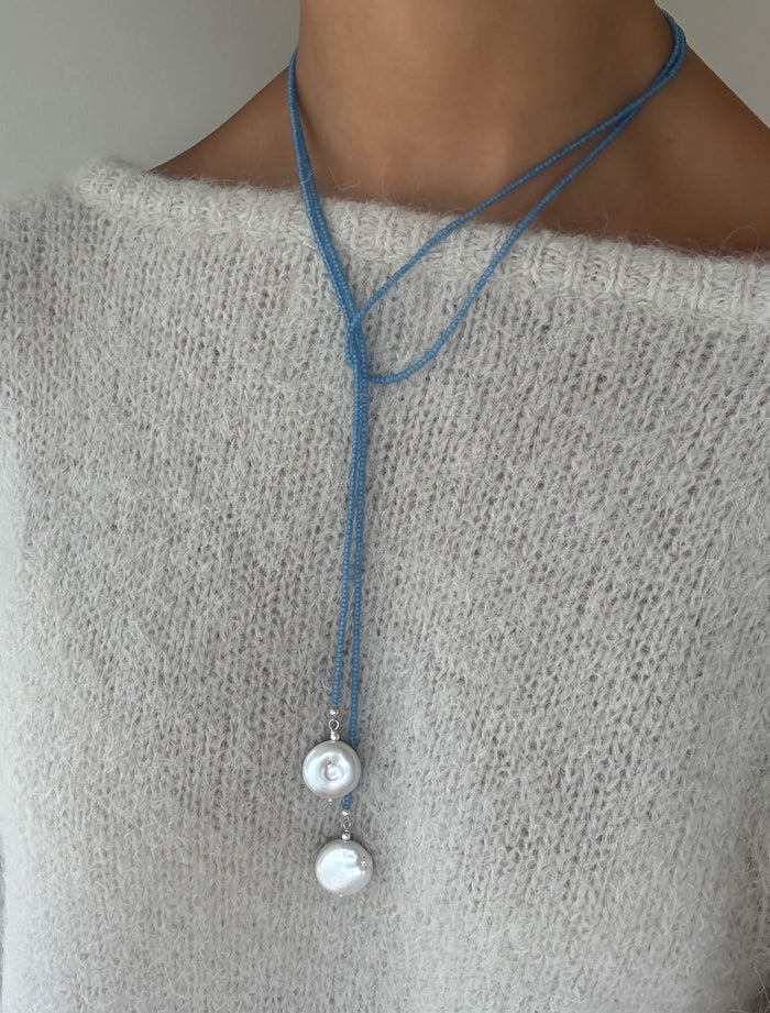 A person wears a soft, light beige knit sweater and the elegant Esme Lariat Necklace by LLUME Jewelry—a double-looped blue cord necklace with two large, shiny white pearls hanging at different lengths.