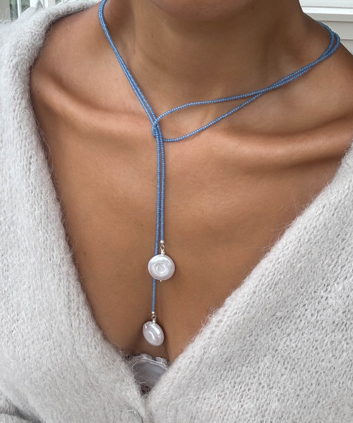 A person wearing a soft, light sweater and the elegant Esme Lariat Necklace by LLUME Jewelry, featuring two white pearl-like pendants—one rose-shaped—draped delicately over their chest.