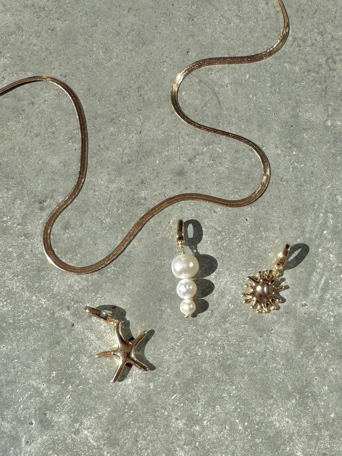 The LLUME Jewelry Every Occasion Set features a gold snake chain necklace on a gray textured surface, accompanied by three interchangeable charms: a starfish, white pearl cluster, and sunburst design.