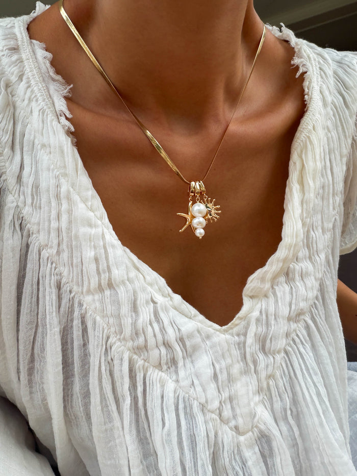 A person wearing a textured white blouse and LLUME Jewelry's Every Occasion Set—a gold snake chain charm necklace with pearl and coral-shaped pendants.