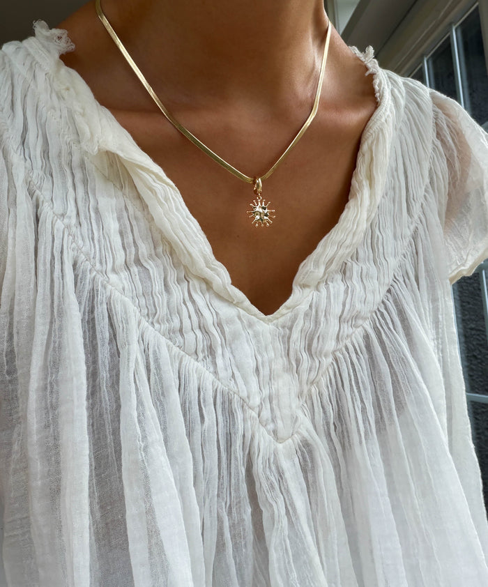 Someone is standing near a window in natural light, wearing a textured white blouse and the LLUME Jewelry Every Occasion Set—a gold snake chain necklace with a sun-shaped pendant.