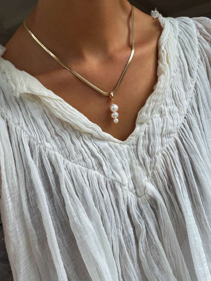 Wearing a crinkled white v-neck blouse, a person accessorizes with the LLUME Jewelry Every Occasion Set—a gold snake chain necklace adorned with four dangling white pearls.