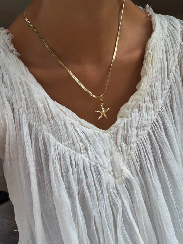 A person wearing a white, textured V-neck blouse and the Every Occasion Set by LLUME Jewelry, featuring a gold charm necklace with a starfish pendant.