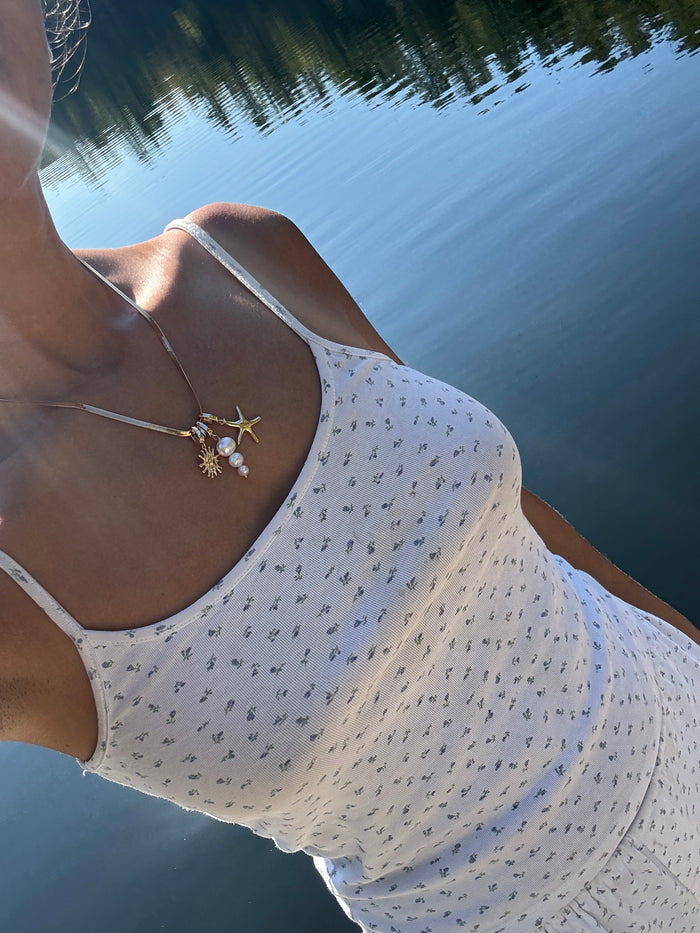 Near a calm lake, a person wears the LLUME Jewelry Every Occasion Set—gold, starfish-themed charm necklaces including a delicate snake chain. They pair it with a white patterned tank; trees and blue sky reflect in the water. Their face is partly out of frame.