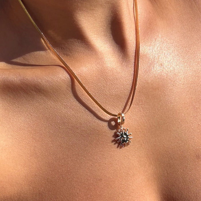 A close-up of LLUME Jewelry's Every Occasion Set shows a gold sun-shaped charm necklace with a snake chain resting on bare skin, its warm shine beautifully highlighted by the sunlight.