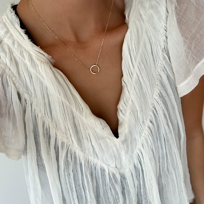 A person wearing a textured white V-neck blouse and LLUME Jewelry's Everything, In Time Necklace—a delicate gold chain with an organic circle pendant. The photo is cropped to show the upper chest and part of the blouse.