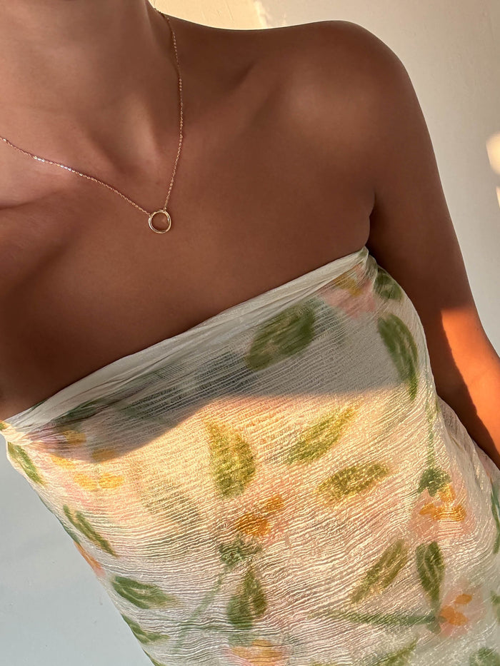 A person wearing a strapless, textured green and yellow floral top and the LLUME Jewelry Everything, In Time Necklace—a gold chain with an organic circle pendant—photographed from the shoulders down in warm natural light.