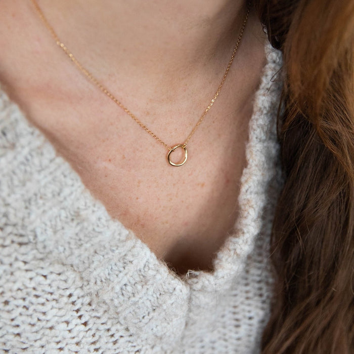 A person wears a light knit sweater and the LLUME Jewelry Everything, In Time Necklace, featuring an organic circle pendant. The image highlights the neck and collarbone, with long brown hair draped on one side.