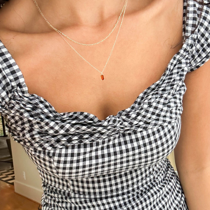 A person wearing a black and white gingham dress with a sweetheart neckline and the Fate Daily Necklace by LLUME Jewelry. The image highlights their upper torso and the necklace’s delicate rectangular pendant.