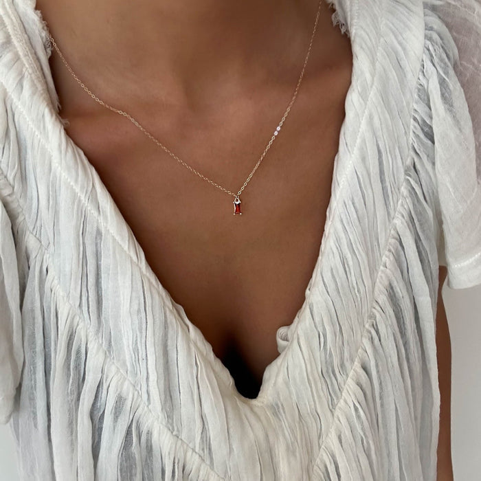 A person wearing a white textured v-neck blouse with pleats and the LLUME Jewelry Fate Necklace, a delicate gold-plated sterling silver piece featuring a small red pendant.