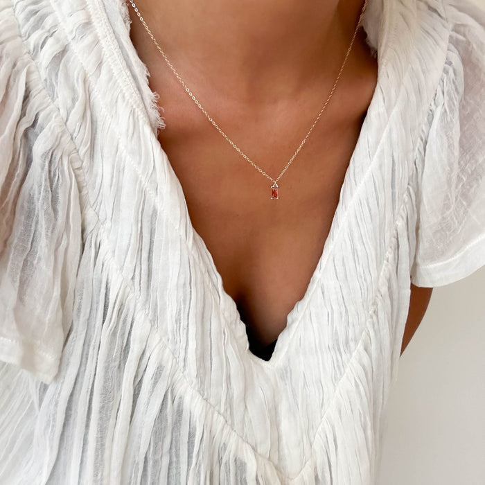 A person wears a white textured V-neck blouse and the LLUME Jewelry Fate Necklace—a delicate gold-plated sterling silver piece with a small red charm. The image highlights the upper chest and neck area.