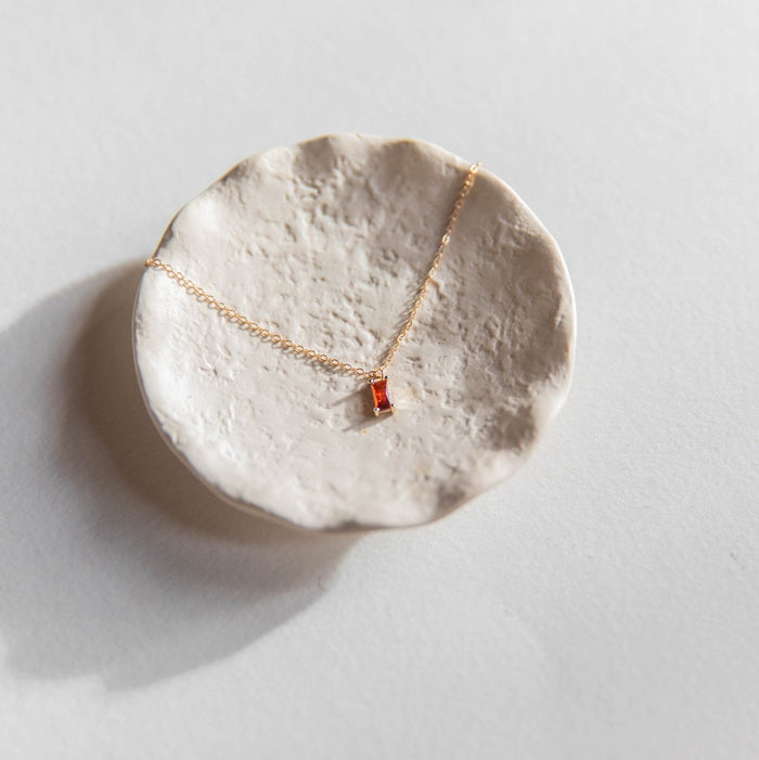 The LLUME Jewelry Fate Necklace, a delicate gold-plated sterling silver chain with a small red rectangular pendant, is displayed on a textured white ceramic dish against a light background.