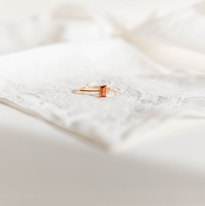 The LLUME Jewelry Fate Ring, crafted in 14k gold-filled metal with a rectangular orange gemstone, is displayed on white embroidered fabric amid a gentle, blurred background.