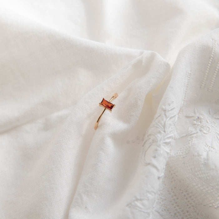 The LLUME Jewelry Fate Ring, crafted in 14k gold-fill and featuring a rectangular red stone, rests on soft embroidered white fabric.