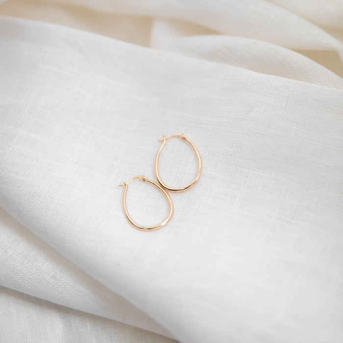 The Fireside Hoops by LLUME Jewelry, delicate 18k gold plated earrings with leverback closures, are displayed on a soft, white textured fabric.