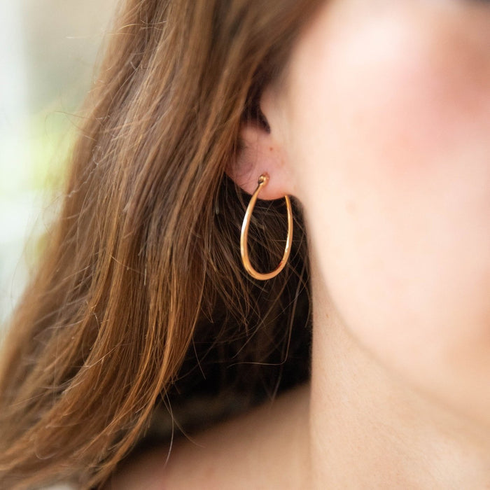 A close-up of a woman's ear shows her wearing LLUME Jewelry's Fireside Hoops, vintage gold earrings with leverback closure. A softly blurred background highlights her brown hair and the elegant design.