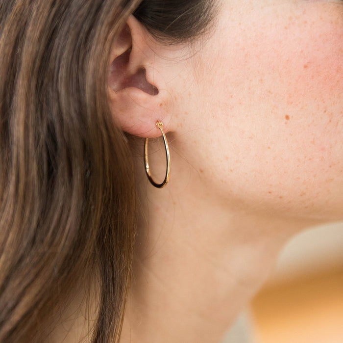 Close-up of a woman's ear with long brown hair, wearing LLUME Jewelry’s Fireside Hoops—simple, shiny 18k gold plated earrings. Freckles are visible on her skin, and the background is softly blurred.