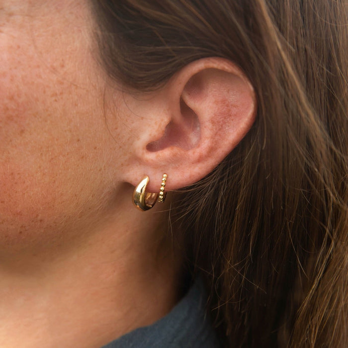 A close-up of a woman with straight brown hair wearing the Go-to Ear Set by LLUME Jewelry: a thick gold hoop and a beaded hoop earring. She is dressed in a dark top.