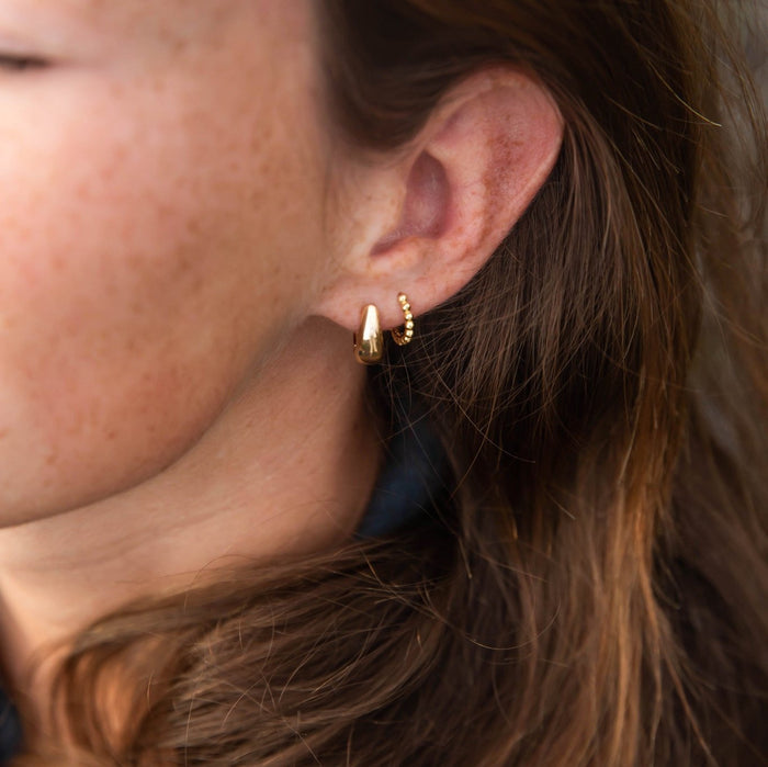 Close-up of a woman's ear wearing the LLUME Jewelry Go-to Ear Set: a smooth teardrop-shaped hoop paired with a small beaded hoop, ideal for stacking. Her brown hair and freckled skin highlight the earrings' elegant design.