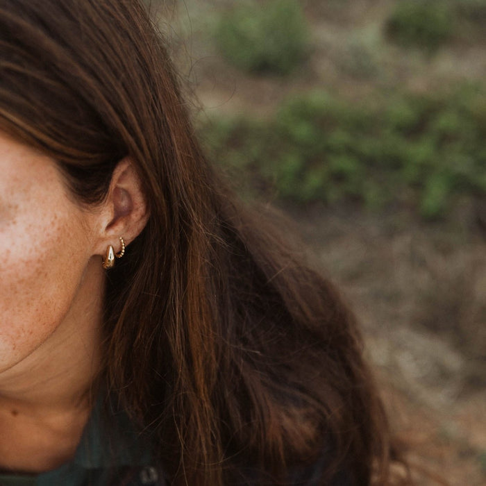 Close-up of a person outdoors with long brown hair and freckled skin, wearing the Go-to Ear Set by LLUME Jewelry. The stylish stacking earrings stand out against a blurred green background.