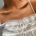 A person wearing the LLUME Jewelry Golden Hour Chain, paired with a white lace-trimmed sleeveless top featuring thin straps and ribbon at the neckline. The photo highlights their neck and upper chest.