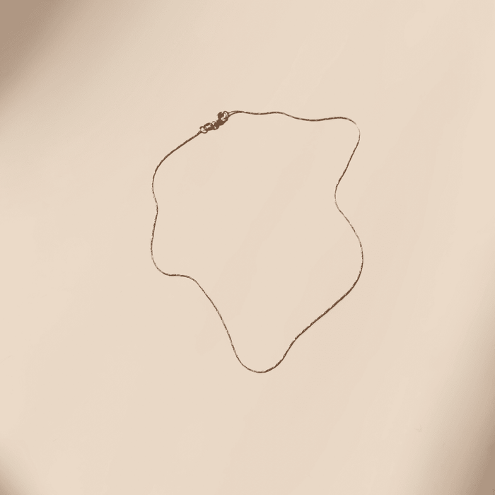 The Golden Hour Chain by LLUME Jewelry is a delicate 18k gold filled necklace with a thin chain and small clasp, displayed in a wavy shape on a plain light beige background.