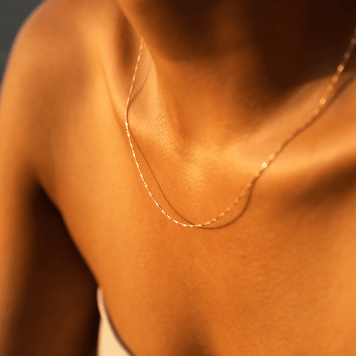 Close-up of a person wearing the LLUME Jewelry Golden Hour Chain, an 18k gold filled necklace that catches warm sunlight on their neck and shoulders. They’re dressed in a light top with thin straps.