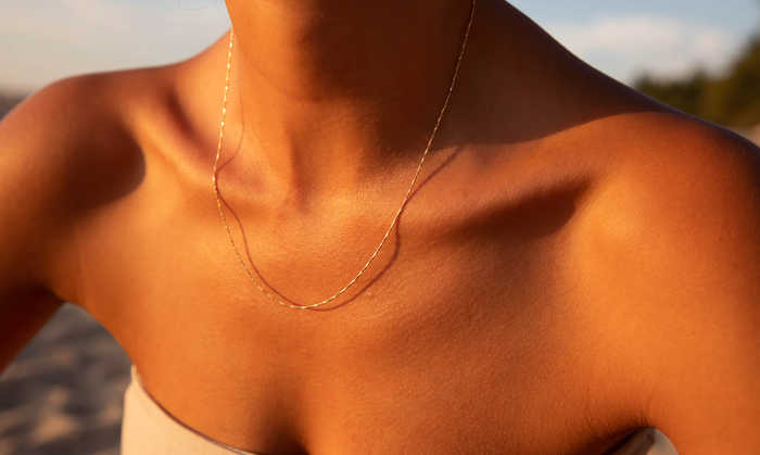 A close-up of a sunlit neck and collarbone adorned with LLUME Jewelry’s Golden Hour Chain, an elegant 18k gold filled necklace, the delicate contours glowing against a softly blurred outdoor background.