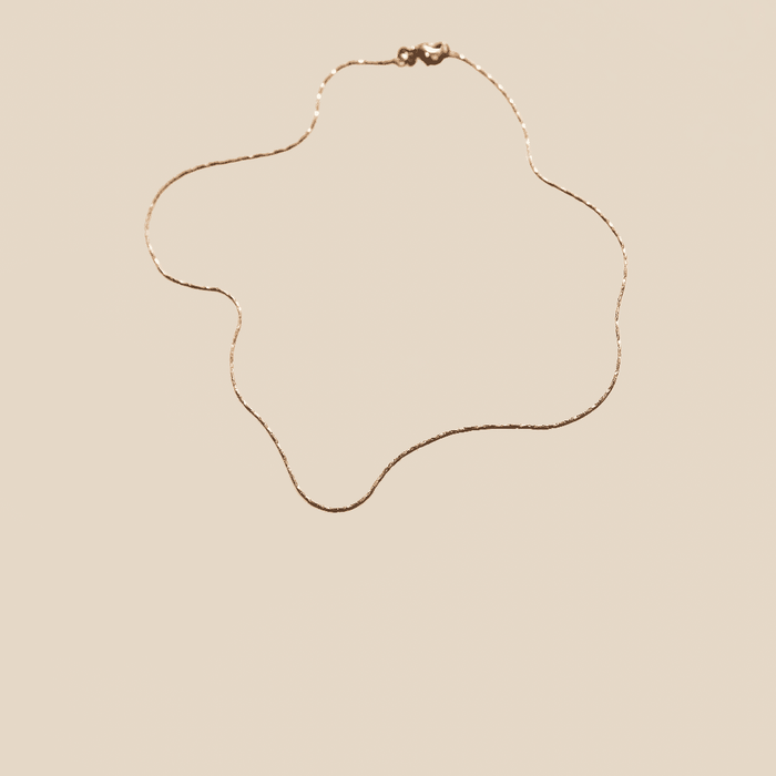 The Golden Hour Chain by LLUME Jewelry, an elegant 18k gold filled necklace, is displayed in an abstract wavy design against a beige background.