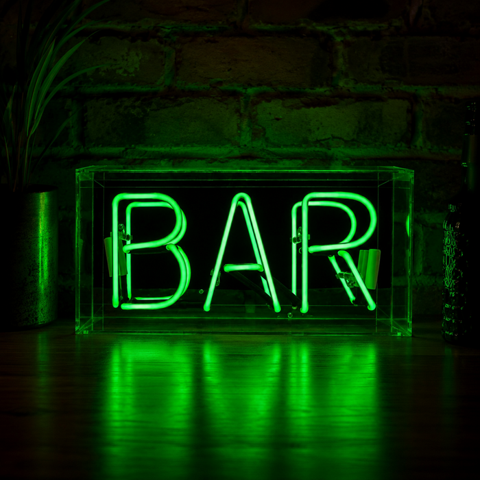 Green BAR neon sign glowing on a wooden surface with dark brick wall background and green light reflections