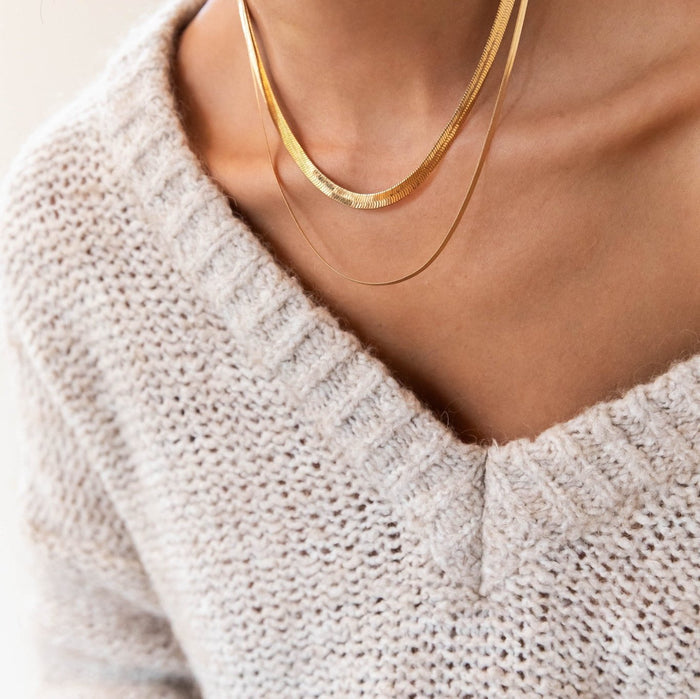Close-up of a person wearing a cream knit V-neck sweater, accessorized with the Hailey Duo layered necklace set by LLUME Jewelry—a snake chain paired with a delicate gold chain.