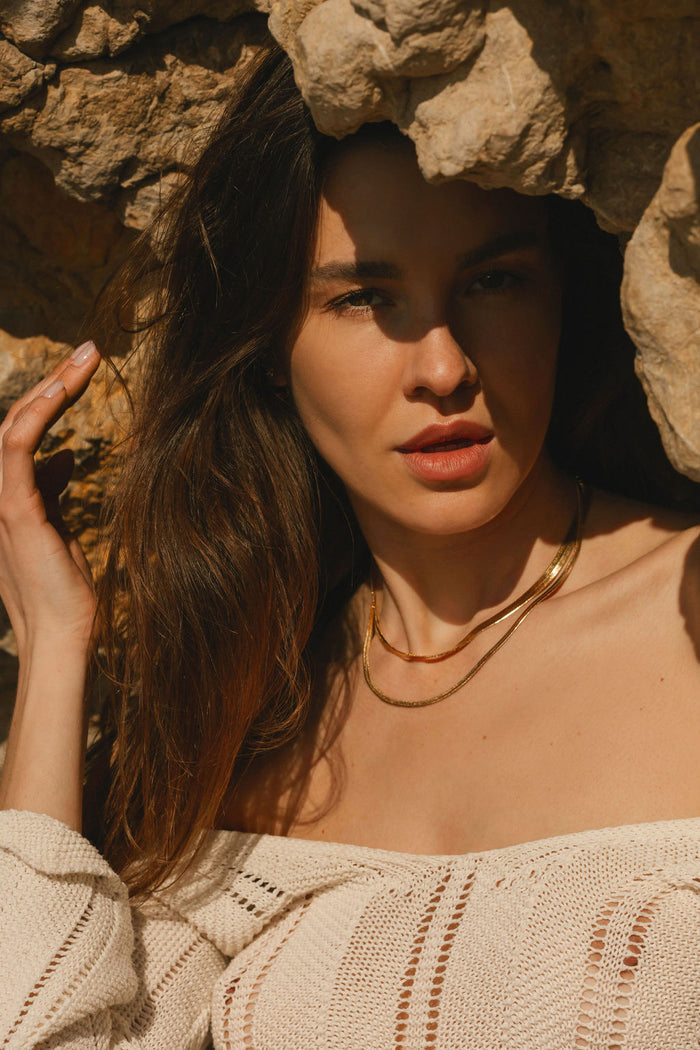 A woman with long brown hair wears a white knit top and the Hailey Duo necklace from LLUME Jewelry as she stands beneath rocky formations, sunlight casting elegant shadows on her face.