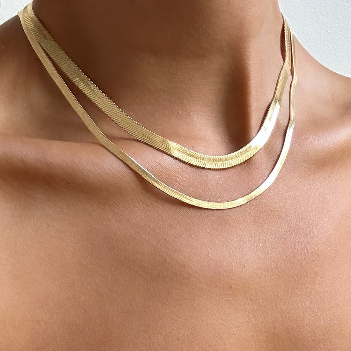 A close-up of a neck and shoulders features the Hailey Duo by LLUME Jewelry—two flat gold herringbone chain necklaces elegantly layered against bare skin.