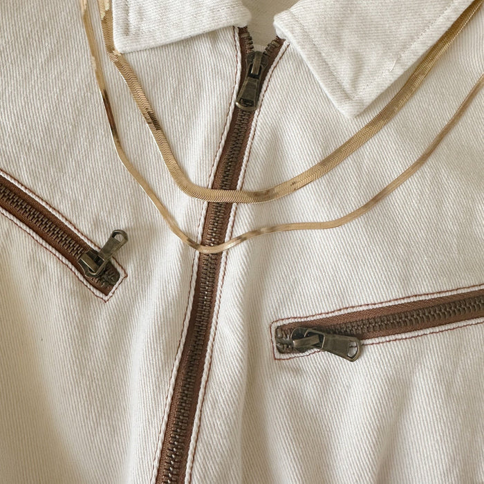 A cream-colored jacket with three brown-trimmed zippers features the Hailey Duo gold snake chain necklace by LLUME Jewelry elegantly draped across the fabric.