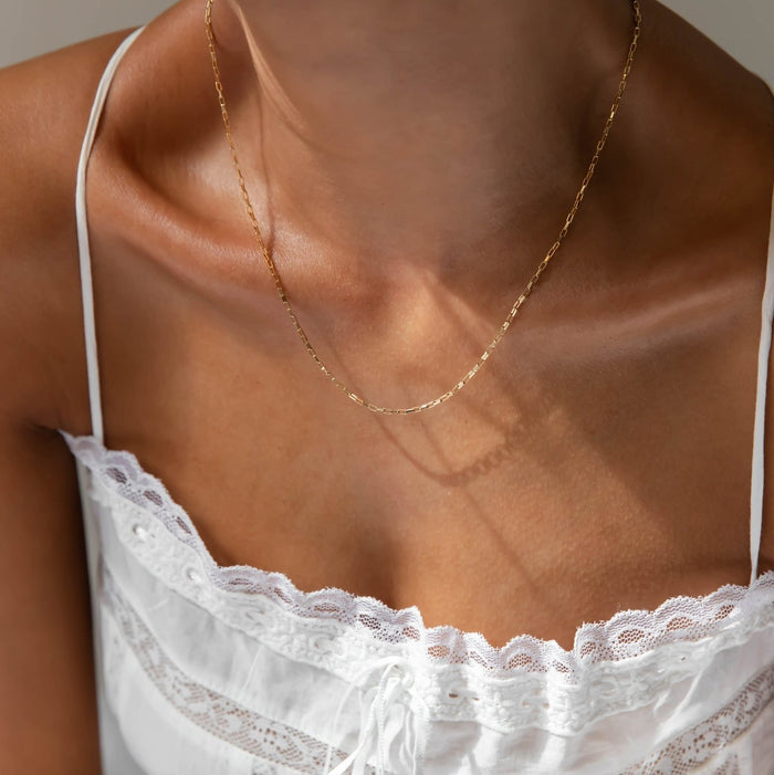 A close-up of a person wearing LLUME Jewelry's Harlow Chain, an 18k gold filled necklace, with a white lace-trimmed camisole, highlighting their neck, shoulders, and upper chest.