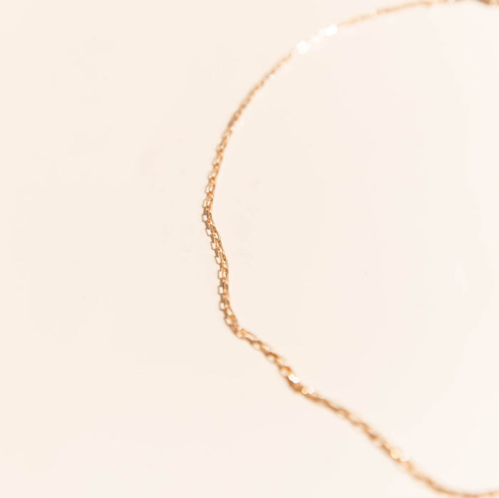 The Harlow Chain by LLUME Jewelry, a delicate 18k gold filled necklace, rests in a gentle curve on a soft beige background with a shallow depth of field softly blurring the chain and backdrop.