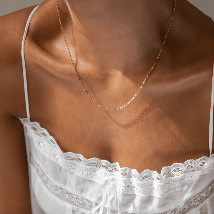 A person wears LLUME Jewelry's Harlow Chain, a delicate 18k gold-filled necklace, paired with a white lace-trimmed camisole, revealing their shoulders and collarbones.