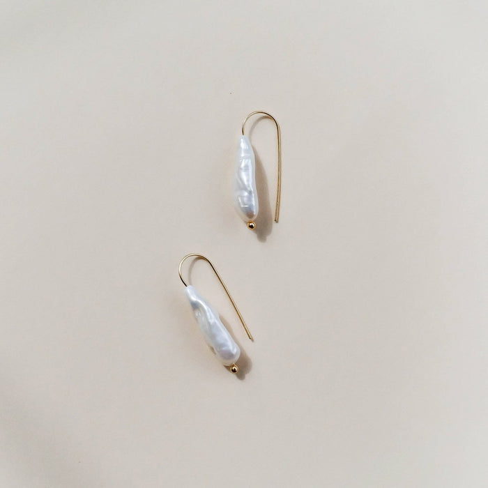 The LLUME Jewelry Here to Stay Earrings feature minimalist, irregularly shaped white freshwater pearls on thin gold plated hooks, showcased against a neutral light beige background.