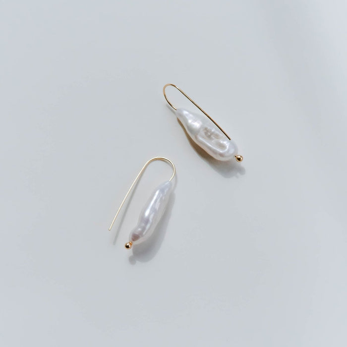 The Here to Stay Earrings by LLUME Jewelry feature gold plating and elongated, irregularly shaped freshwater pearls. Their minimalist design highlights each pearl's unique natural form, set against a smooth, light grey backdrop.