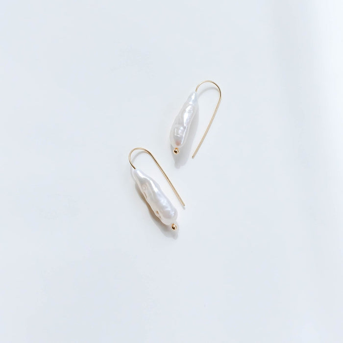 The Here to Stay Earrings by LLUME Jewelry, featuring gold plating and elongated white baroque freshwater pearls, are displayed on a clean white surface, highlighting their timeless elegance.