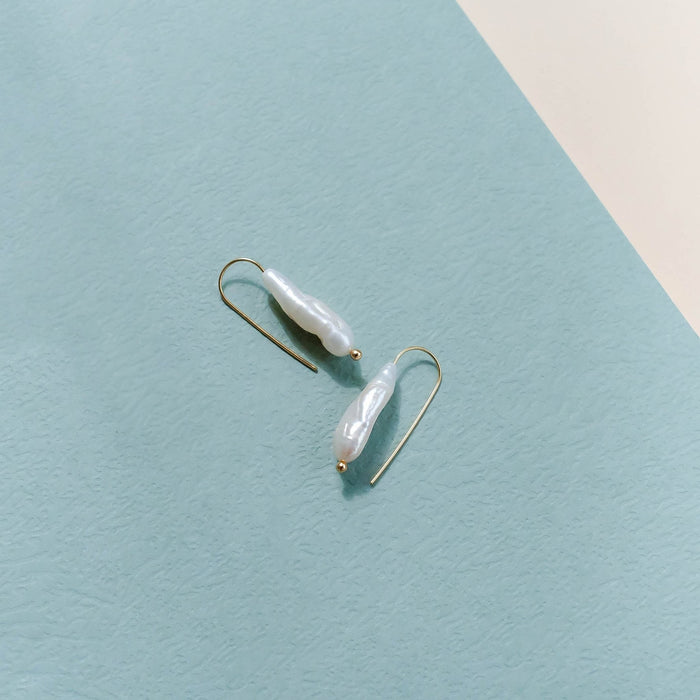 The Here to Stay Earrings by LLUME Jewelry, featuring elongated white freshwater pearls with gold plating, are displayed on a textured light blue surface with a cream corner, highlighting their timeless elegance.
